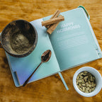 Open book titled 'Happy Hormones' with a bowl of nuts, spoon, and small pot on a wooden surface.